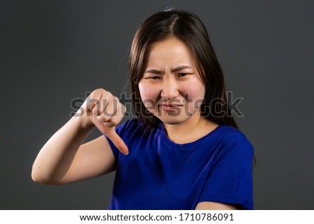Young korean girl standing on grey studio background expressing discontent and showing thumb down gesture at camera. Portrait of asian woman with sign of dislike.