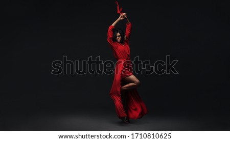 Full length of a young elegant woman dancing contemporary dance. Female in red dress performing strong dance moves over black background.