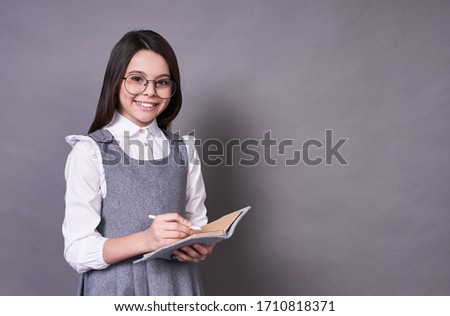 Beautiful young emotional lady, schoolgirl, long-haired brunette in glasses, with a notebook and pen, makes notes, the concept of study,looks at the camera and smiles broadly, on a gray background.