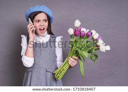 Portrait of a beautiful schoolgirl lady French brunette, in a stylish blue beret hat, in her hands with flowers tulips upset and upset by the phone call,September 1,school, study,on a gray background.