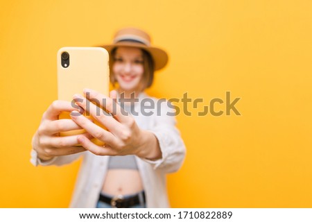 Happy girl tourist in light clothing and hat makes selfie on yellow background, focus on hand with smartphone, close up photo. Isolated. Copy space