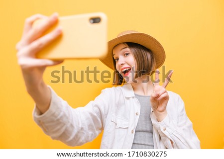 Happy tourist girl in sun hat and summer clothes makes selfie on smartphone on yellow background, looks into camera and shows gesture of peace. Lady taking photo on smartphone camera.