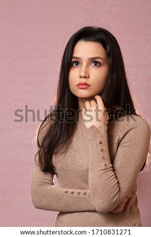 Portrait of a young beautiful Asian brunette in a light brown sweater thinking about something on a pink isolated background with copyspace