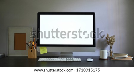 Photo of Workspace blank screen computer monitor putting on working desk and surrounded by picture frame, pencil holder, stack of books, wireless mouse, keyboard, coffee cup and wild grass in vase.