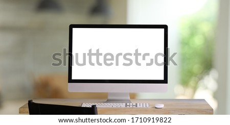 Computer monitor with white blank screen putting on workspace wooden desk with keyboard and mouse over blurred living room as background.