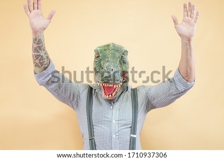 Senior man wearing t-rex dinosaur mask. Crazy man having fun celebrating a party. Absurd and surreal funny concept. Yellow background - Image