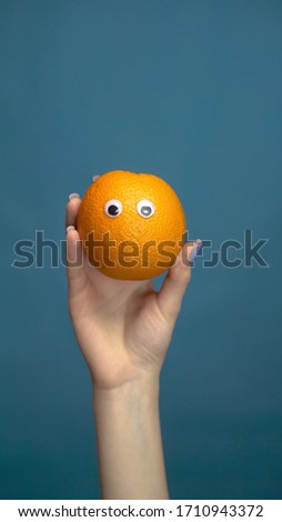 Orange with eyes in a woman hand close up. Orange looks around on a blue background. Vertical photo.