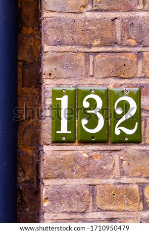House number 132 as digits on green tile screwed onto a brick wall