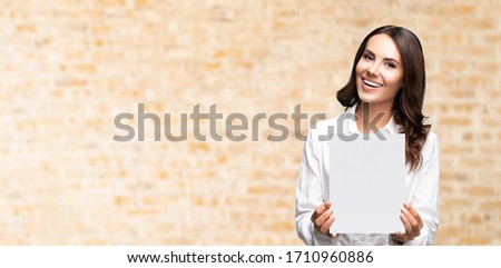 Portrait of happy smiling brunette businesswoman showing blank signboard with copy space area for some slogan or text, over loft style wall background. Success in business concept. 