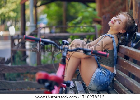 Teenager girl sits with raised legs on a park bench and smiles. In front of her is a bicycle.