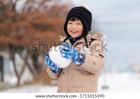 Portrait of happy little Asian boy holding a snow ball, having fun in a winter park.