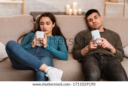 Boredom Concept. Young couple using smart phones, sitting on couch at home during self isolation