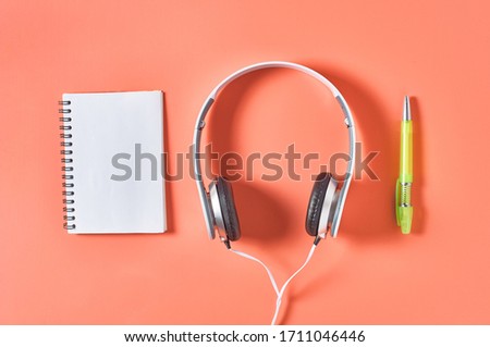 Modern white headphones with cable near blank paper notebook and pen on orange background. Music concept. Copy space. Top view
