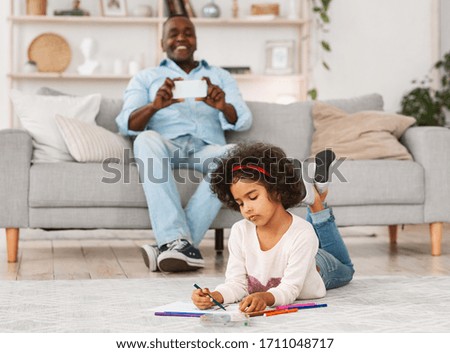 Happy African American granddad taking picture of his cute granddaughter drawing on floor