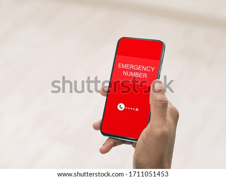 Top view over shoulder of hand holding smart phone with emergency number on red screen over blurred background Royalty-Free Stock Photo #1711051453