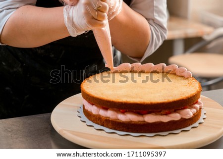 Confectioner woman creating a cream berry cake with strawberry. Cooking process photo with hands. Proffesional chef at work on bakery kitchen.