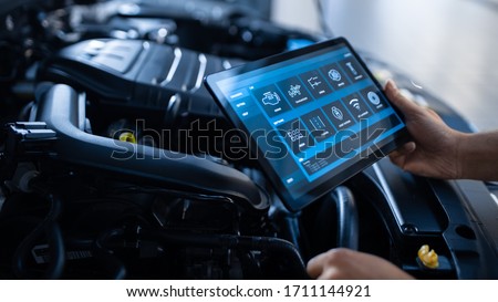 Car Service Manager or Mechanic Uses a Tablet Computer with a Futuristic Interactive Diagnostics Software. Specialist Inspecting the Vehicle in Order to Find Broken Components In the Engine Bay. Royalty-Free Stock Photo #1711144921