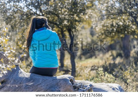 brunette girl with long hair dressed in blue sitting on a dry log in the field talking to her cell phone