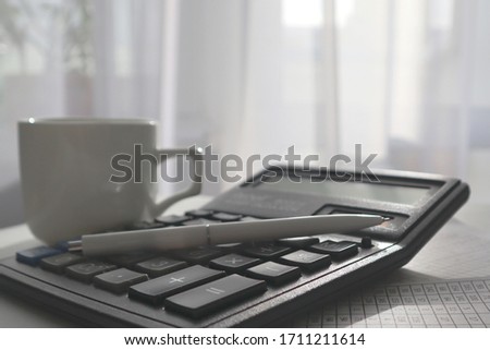 A white pen lies on a black calculator. A white cup is standing nearby. Business concept.
