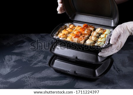 Cook wearing disposable gloves is packing sushi rolls into the black container. Food delivery concept