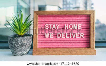 COVID-19 "STAY HOME WE DELIVER" Coronavirus social distancing restaurant business message sign with text offering online delivery to home, staying inside. Pink felt board with plant.