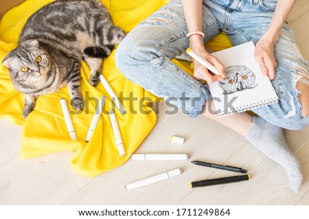 Drawing at home. A girl in blue jeans sketch a gray fold cat with yellow eyes. Nearby are markers, a yellow plaid. The cat is sitting near the girl. View from above, flat lay.