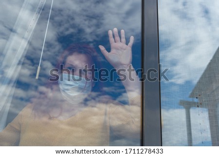 Redhead woman with yellow sweater and mask to prevent coronavirus looking out the window with the reflection of the clouds fading