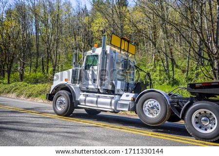 Powerful big rig semi truck with oversize load yellow sign on the roof transporting cargo on the step down semi trailer running uphill on the winding mountain road with forest trees on the side