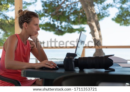 Young female photographer checking and editing photos on her laptop computer, sitting at a desk outside in summer resort.