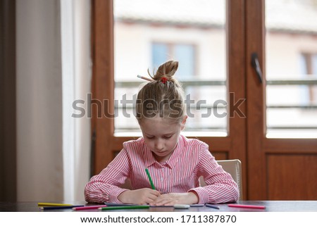 Close up of cute blond hair school girl, sitting at the desk with colored pencils and paper, drawing, closed at home
