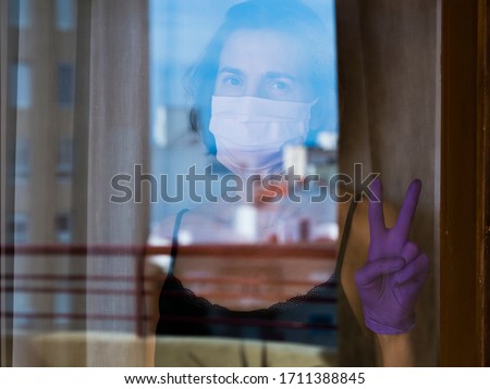 Woman with medical mask and medical gloves, behind her home glass window, making the victory sign hand, during the coronavirus quarantine