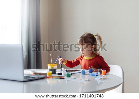 Young cute girl in orange dress spending a time drawing using online lessons on laptop. Distance learning online education,  girl in quarantine stay at home in self isolation.