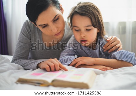 Mother and her daughter reading from bible and praying in their knees near the bed Royalty-Free Stock Photo #1711425586