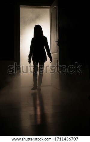 A silhouette of a little girl emerges from a luminous room with smoke in the dark. The concept of children's fears and mysticism.