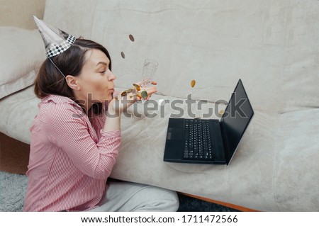 online greeting from video link. young woman in  festive hat makes a salute of Golden confetti to laptop screen at home. Remote communication during the quarantine period.