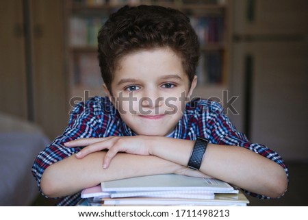 Teenager boy sits at a table and folded his hands on a stack of books. Smiling and looking at camera. Training at home on coronavirus quarantine. Selective focus. Blur background.
