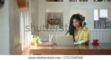 Photo of woman sitting and taking note while sitting in front her computer laptop that putting on wooden working desk and surrounded by coffee cup, pencil holder, potted plant and stack of books