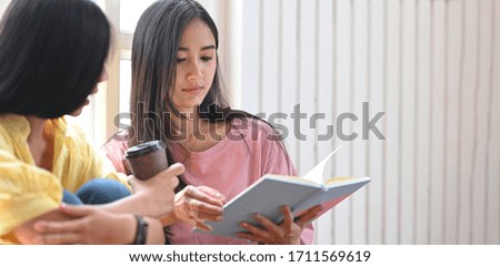 Photo of young sisters relaxing/reading a notebook while sitting together at the wooden floor over comfortable living room windows as background.