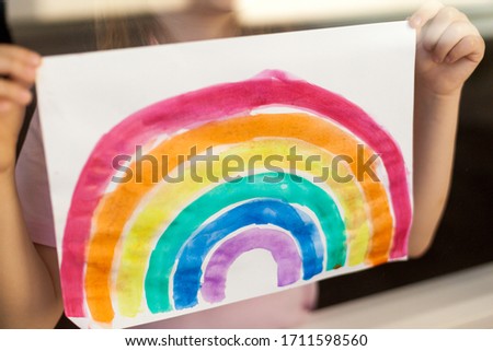 a girl holds a drawing of a rainbow near the window