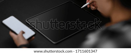 Cropped shot of female entrepreneur working at dark modern office desk with mock-up smartphone and digital tablet on black table