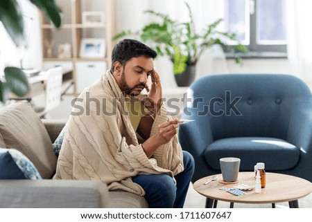 people, health and fever concept - sick indian man in blanket measuring temperature by thermometer at home Royalty-Free Stock Photo #1711607647