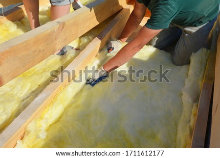 Roofing construction: building contractors in protective gloves are installing glass wool sheets, batts on the rooftop below trusses for thermal insulation of the rooftop. Royalty-Free Stock Photo #1711612177