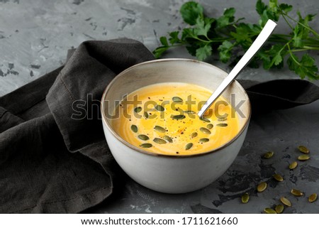 Pumpkin soup in deep bowl with seeds and spoon served with napkin and fresh green on grey background. 