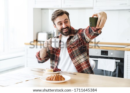 Portrait of smiling bearded man taking selfie on cellphone and drinking coffee while having breakfast at home