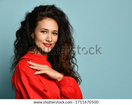Portrait in profile o smiling adult woman with long curly hair posing in red sweater with her hand wt her shoulder over light blue background with free text copy space