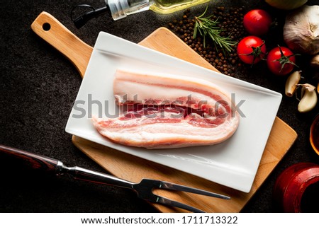 raw pork belly with garlics, tomatoes, rosemarry,Olive oil, pepper, bbq sauce and carving fork on stone black background, dark picture style.
