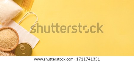 Dry goods, delivery, stockpiling, food supplies for staying home concept, donation, volunteer. Craft paper shopping bag, preserves, pasta, oatmeal, rice, sugar, on yellow background, copyspace, banner Royalty-Free Stock Photo #1711741351