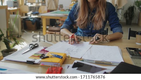 Inspired woman fashion designer creating sketches with brush at workplace, needlewoman works in studio workshop on new clothes collection ideas, making ethnic cross-stitch embroidery design pattern