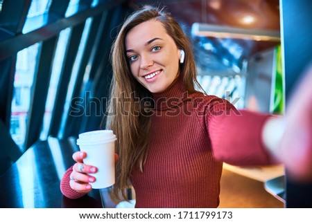 Casual happy young woman with wireless headphones and paper coffee cup takes a selfie photo on the phone while relaxing in a cafe. Modern people with digital mobility lifestyle