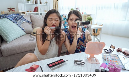 two cute girl friends enjoying free time in modern light white interior living room at home. ladies making funny face looking at camera posing with cosmetics brushes. female makeup taking picture